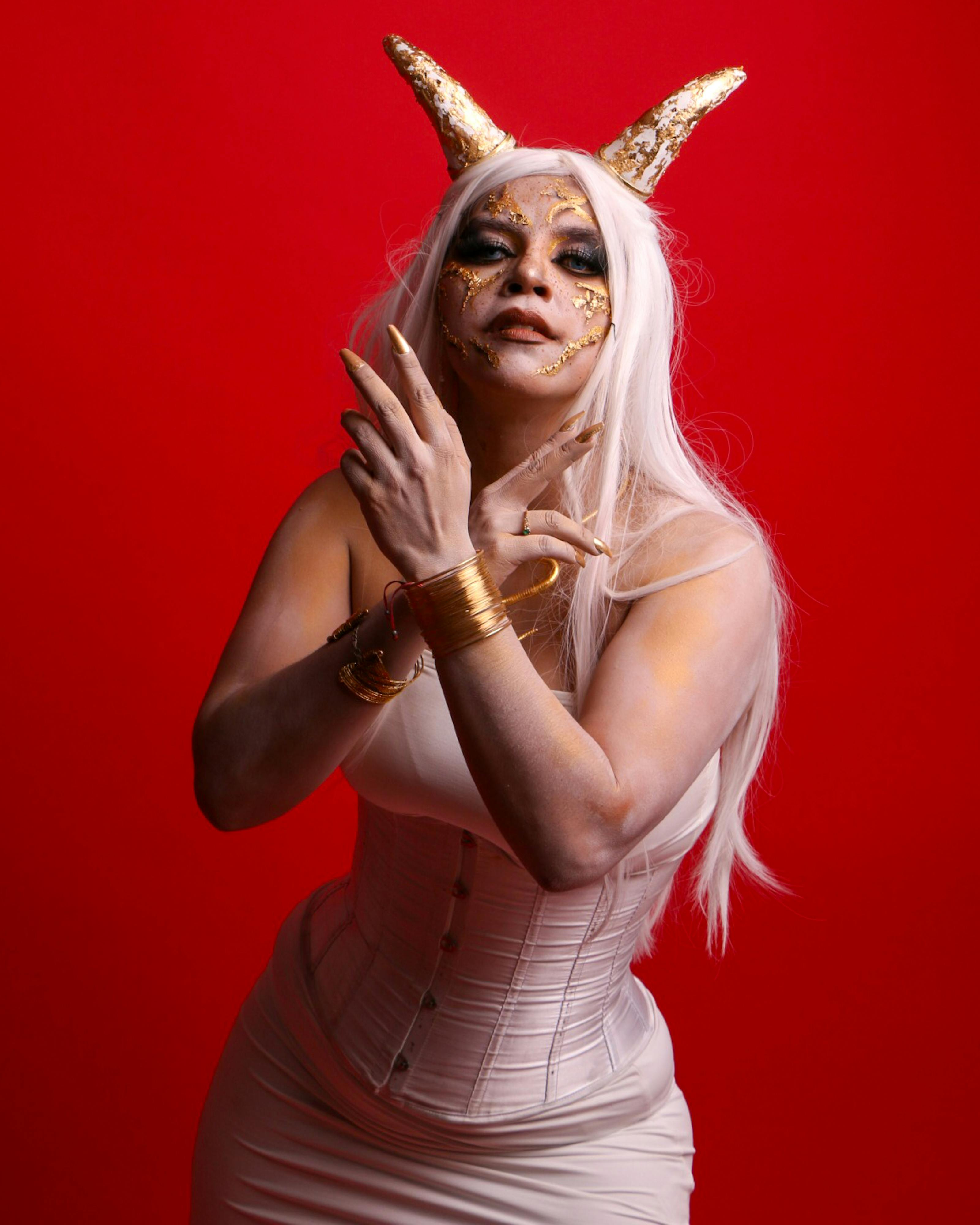 Creative portrait of a woman with gold horns and detailed face art against a red background.