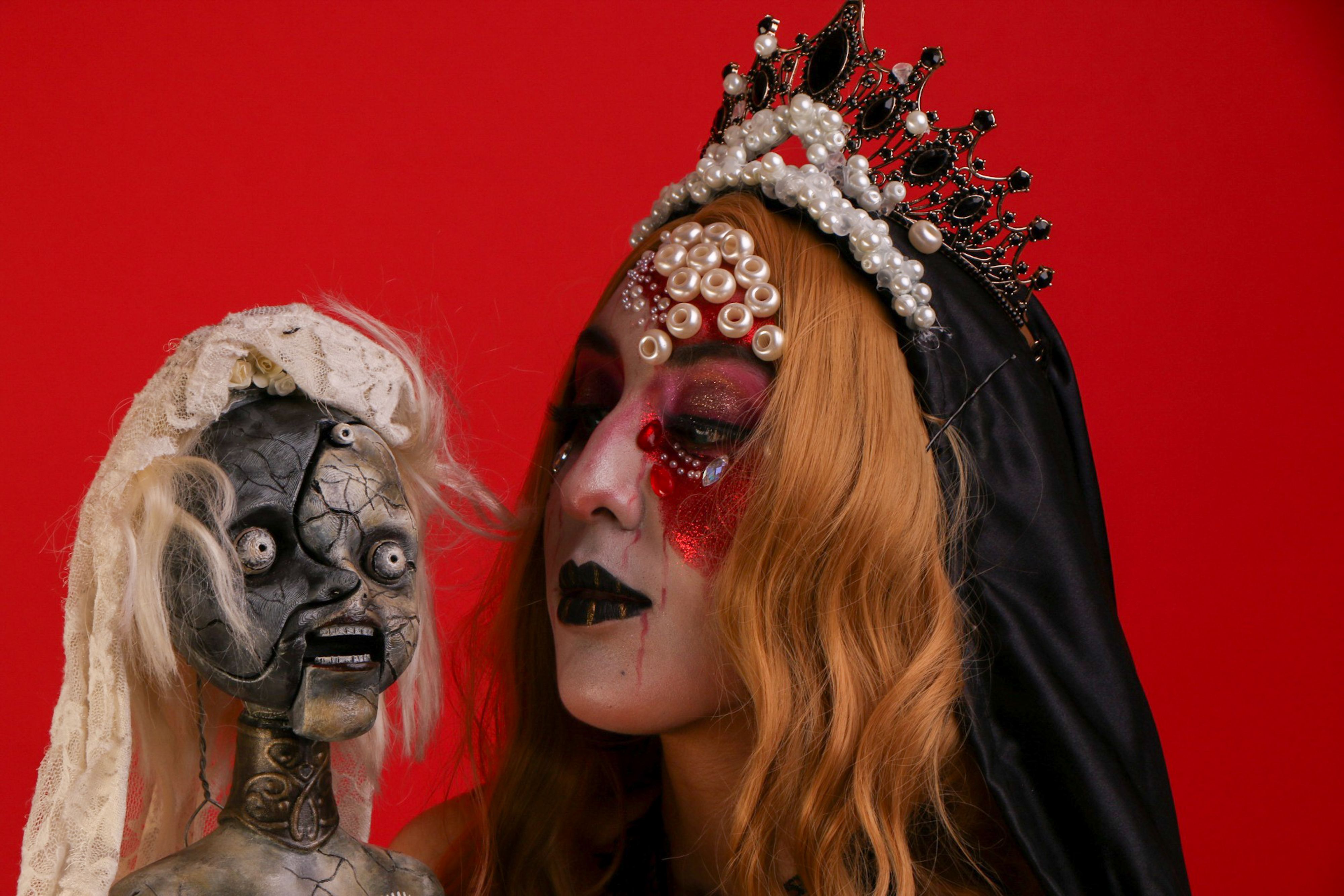 Gothic woman in elaborate makeup and crown holding an eerie doll against a vibrant red backdrop.