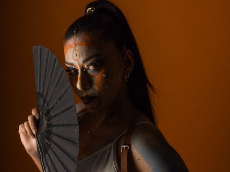 Striking portrait of a woman with artistic makeup holding a fan against an orange background.
