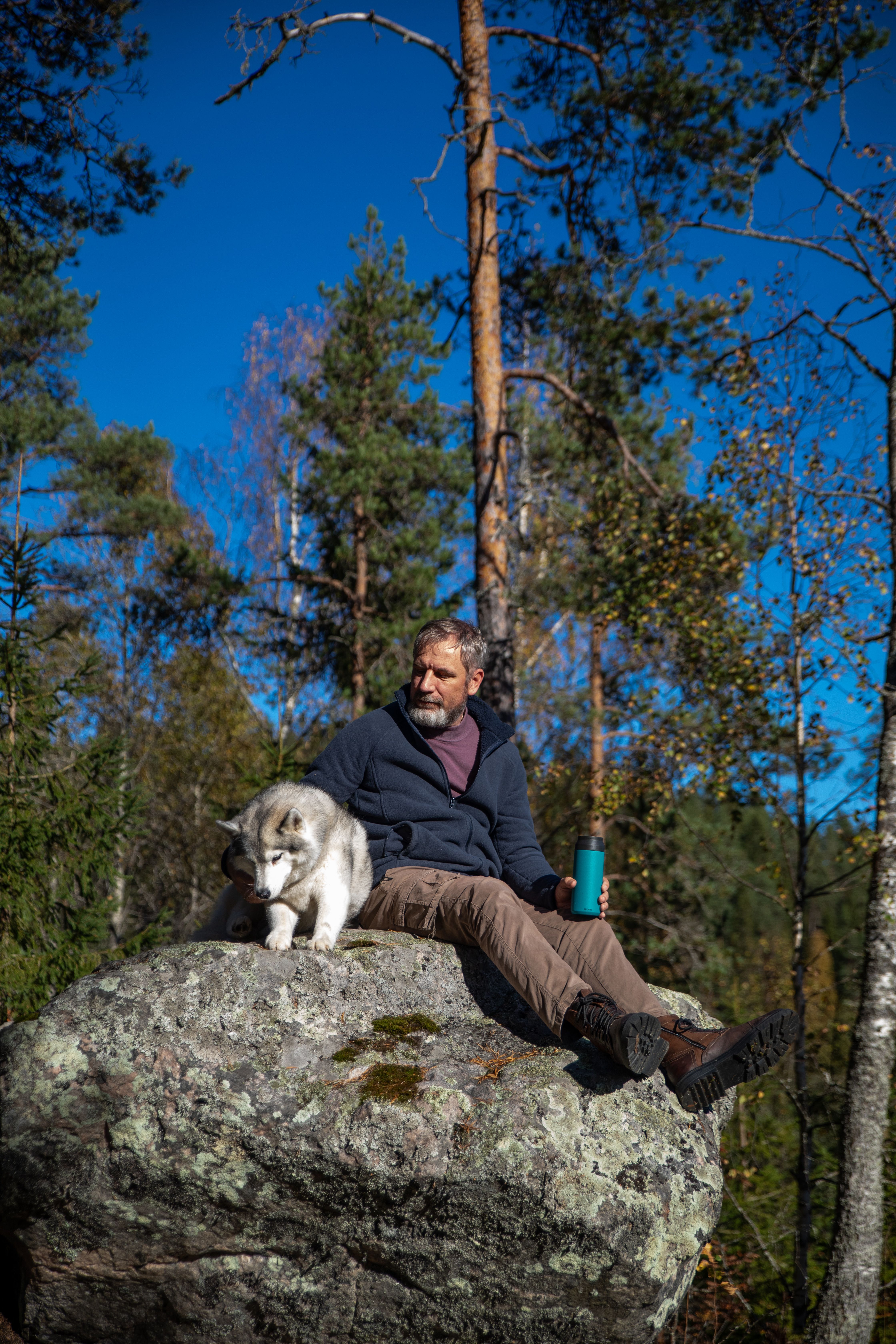 Man enjoying nature on a rock with his husky in a forest setting under a bright sky.