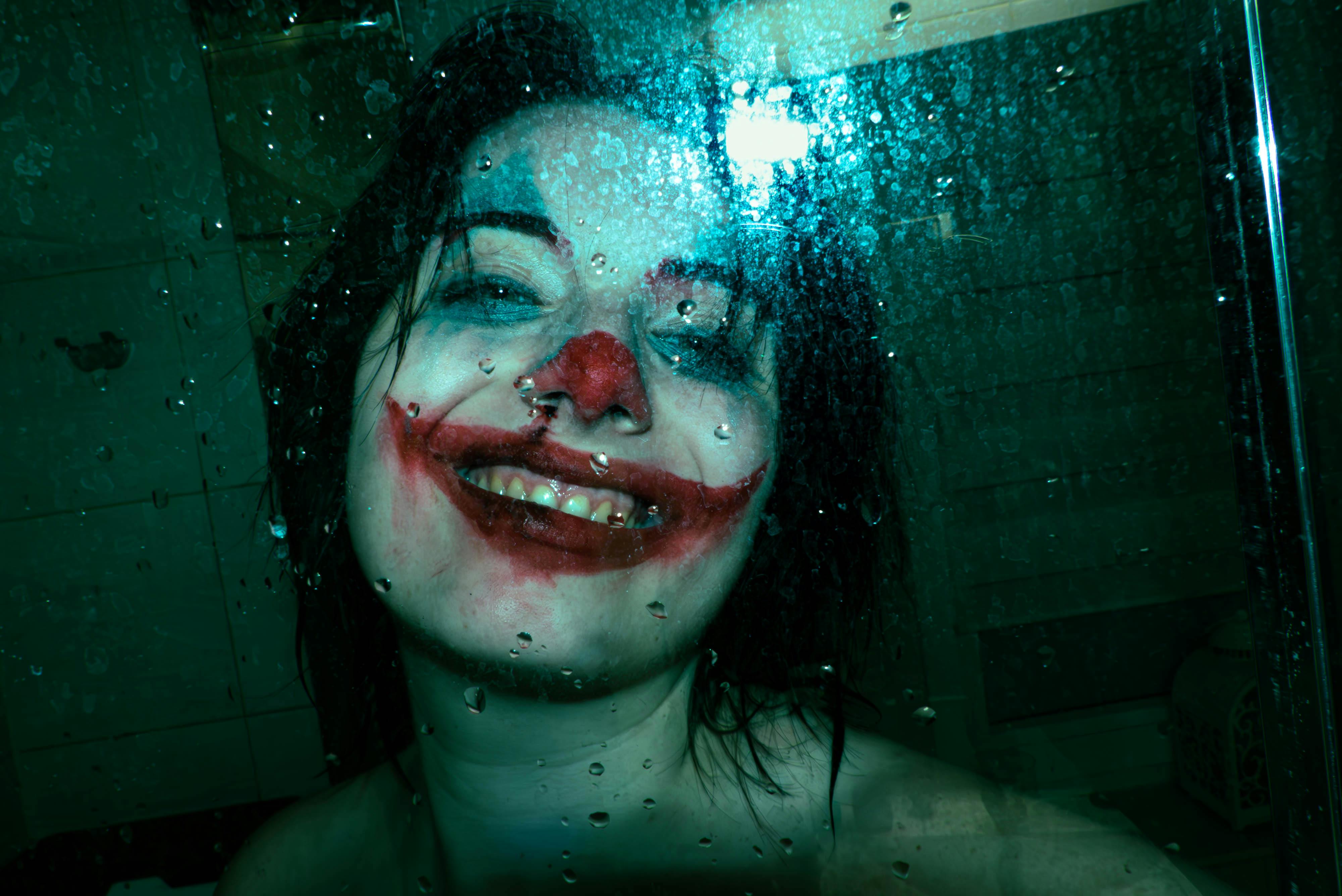A person with smeared Joker makeup smiles eerily behind a wet glass surface.