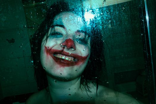 A person with smeared Joker makeup smiles eerily behind a wet glass surface.