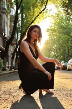 Stylish woman in black dress crouching on sunlit street, urban backdrop.