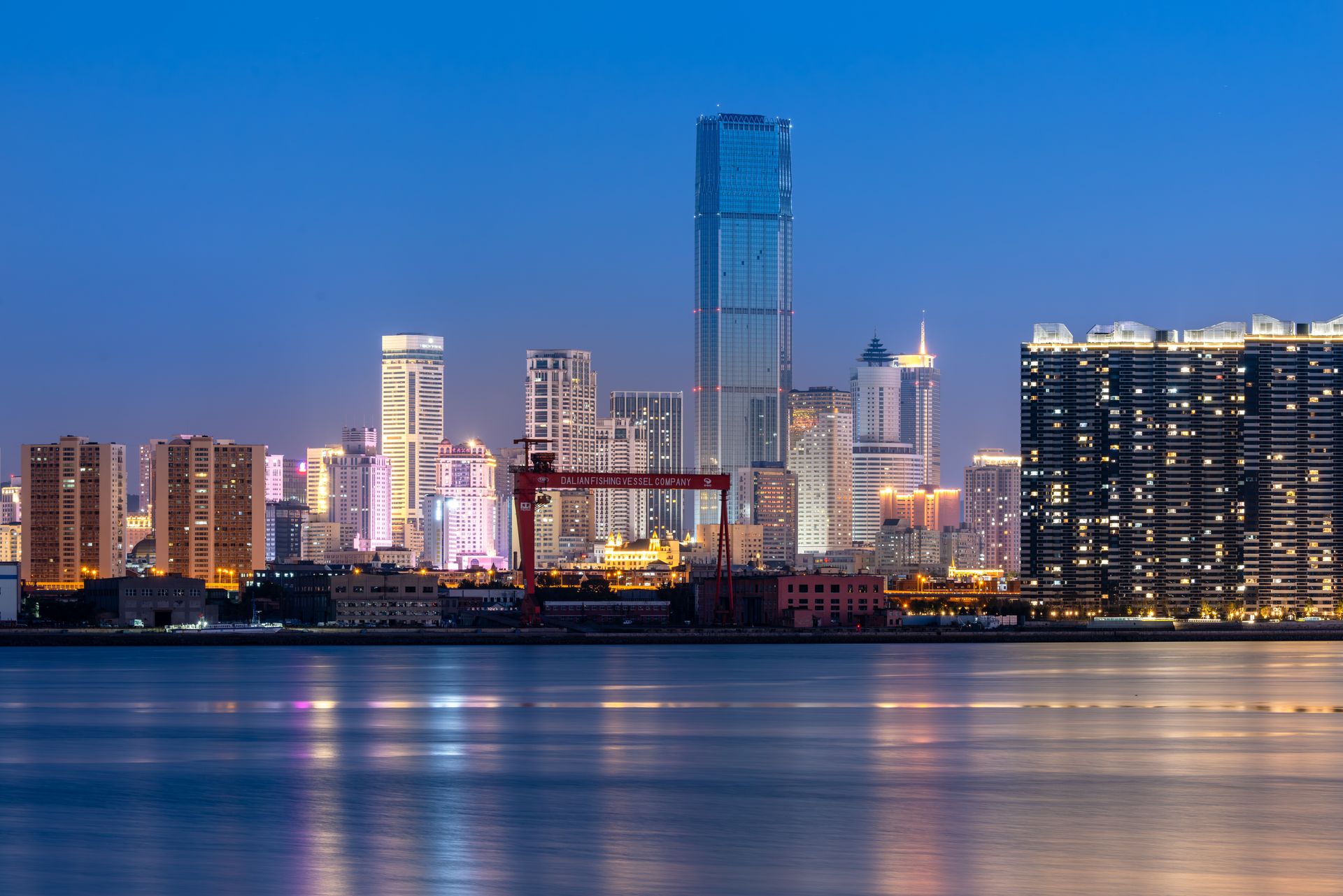 Illuminated city skyline reflecting on calm water at twilight.