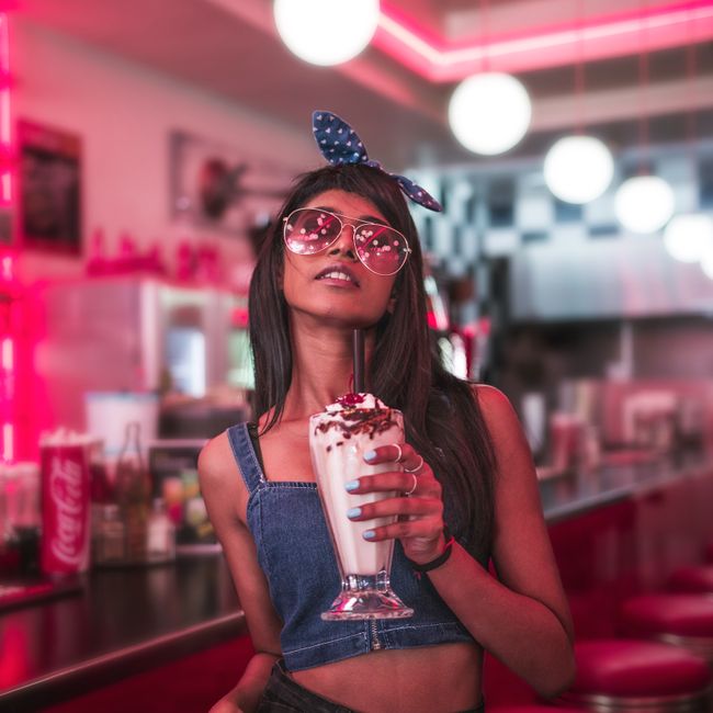 Young woman in fashionable attire enjoying a milkshake at a retro diner with vibrant lighting.