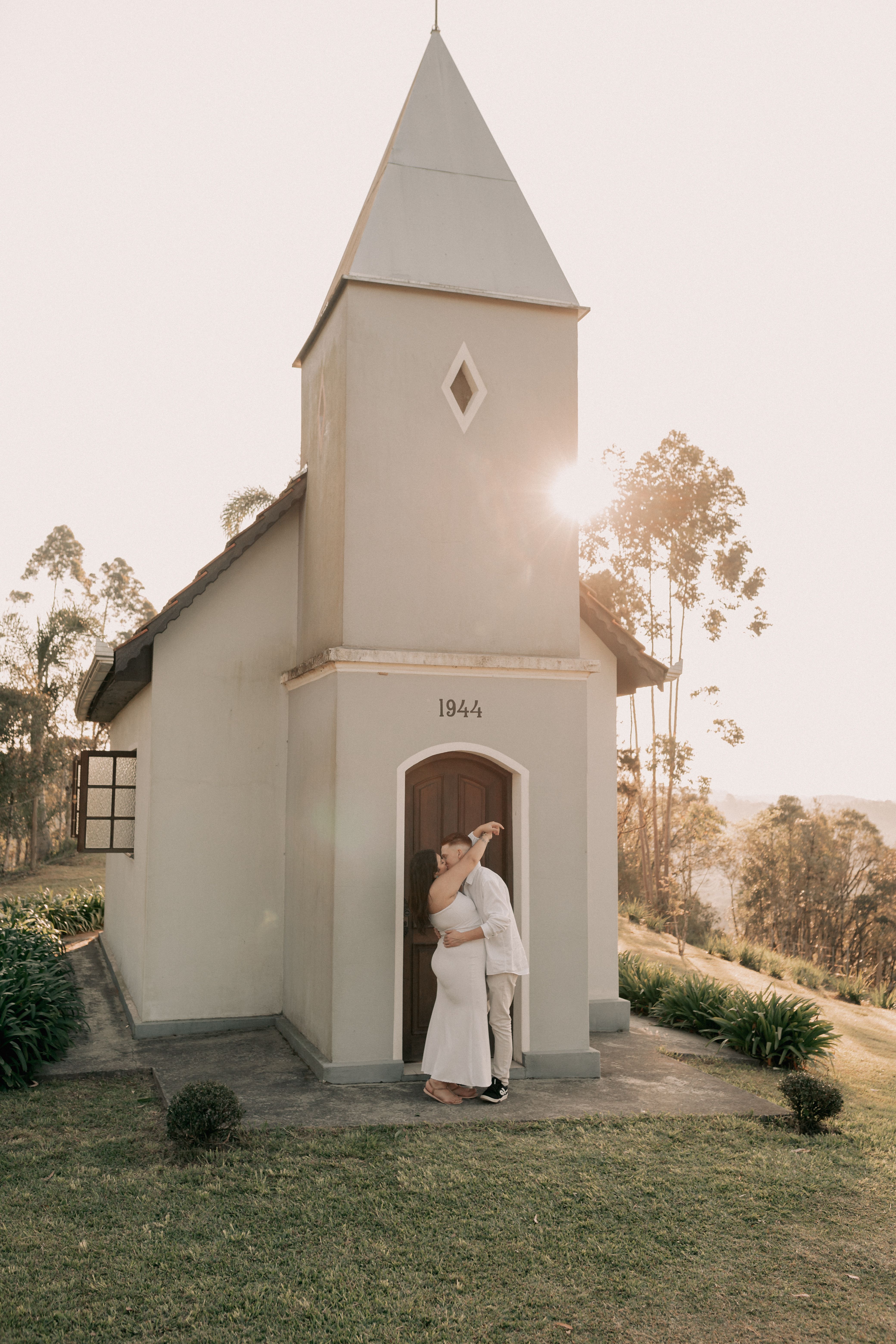 A couple embraces in front of a quaint historic chapel, glowing in the sunset light.