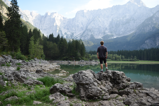 A lone man stands on rocks overlooking a serene alpine lake and mountains.