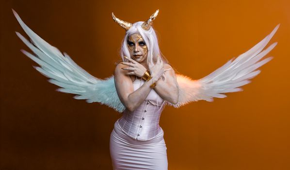 Artistic portrait of a woman with white wings and golden horns on an orange background.