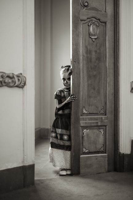 A young girl with Catrina makeup peeks through a door in Mexico City, embodying Day of the Dead spirit.