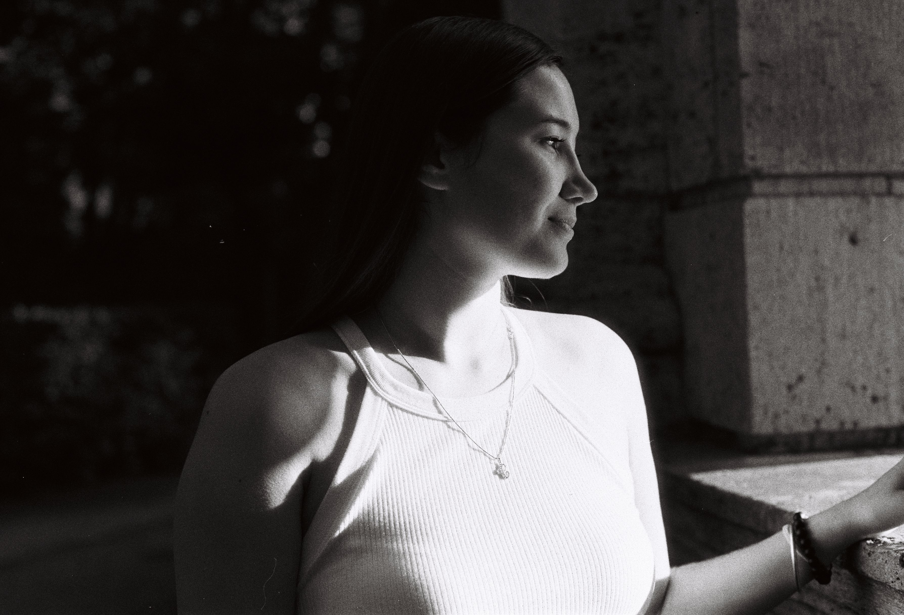 Black and white portrait capturing a woman standing by a wall in sunlight.