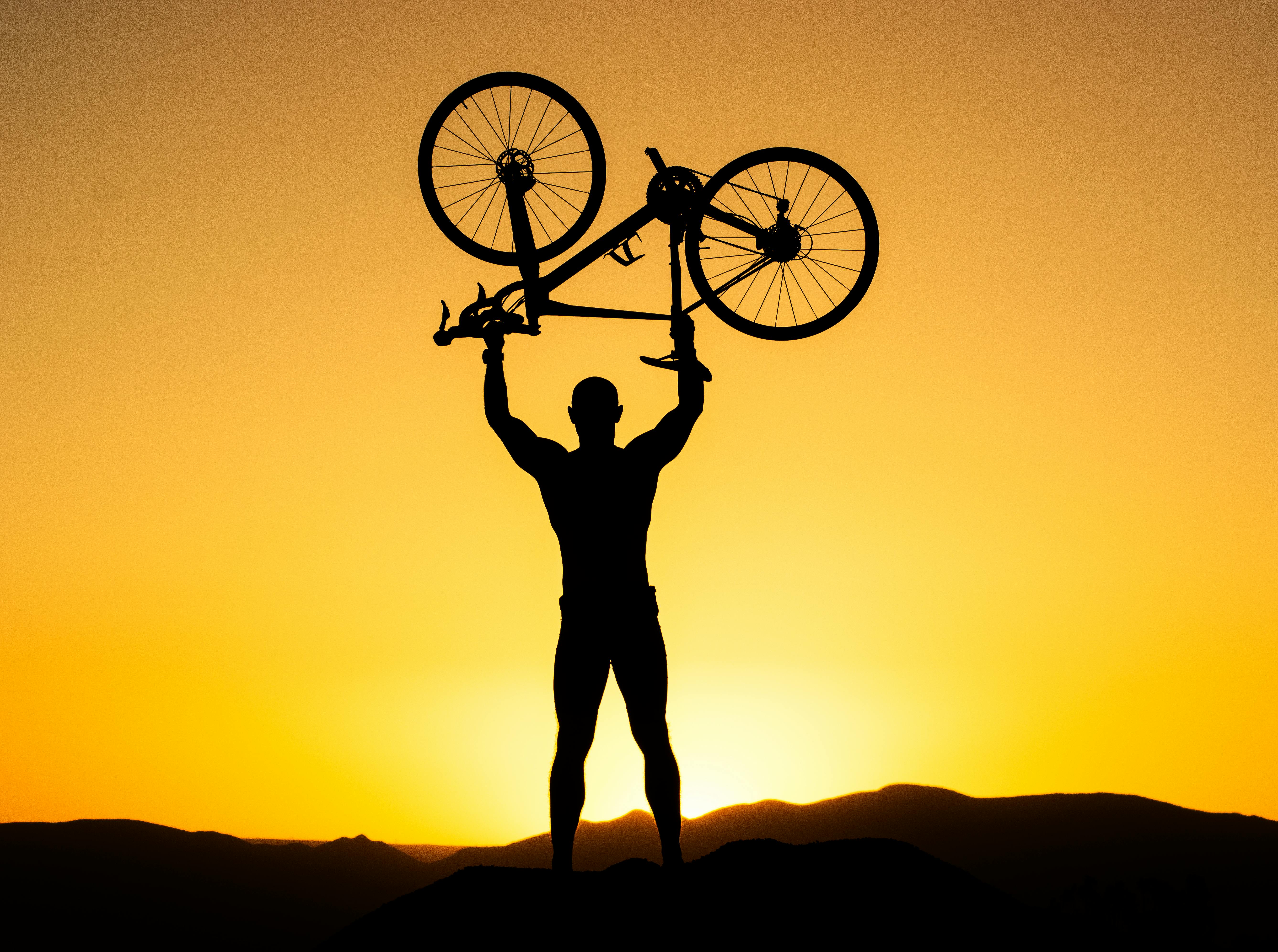 Silhouette of a strong cyclist holding a bike over their head against a stunning sunset.