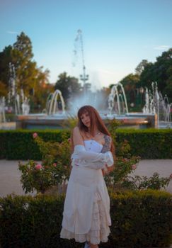 A woman in a white dress poses gracefully in front of a scenic garden with fountains.
