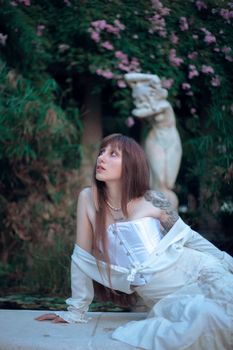 A woman in a white dress poses near a classical garden statue, evoking elegance.