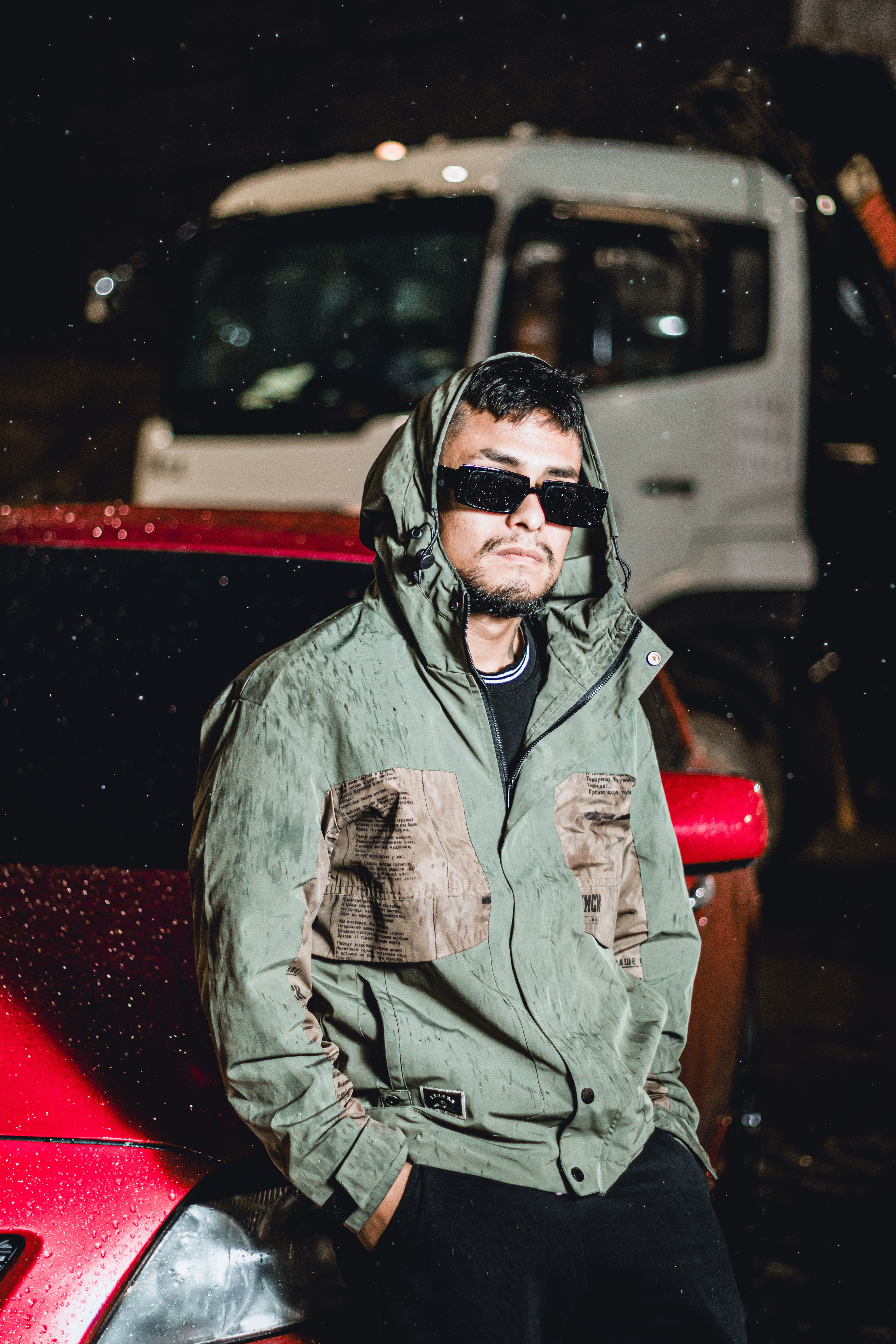 Fashionable young man in a hoodie and sunglasses leaning on a car at night with raindrops.