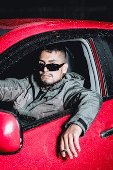 Stylish man in sunglasses leaning out of a red car window during rain. Night scene with moody ambiance.