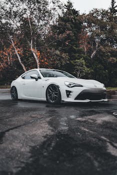 Stylish white sports car parked outdoors on a rainy day, surrounded by trees.