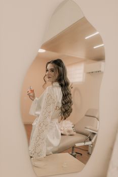 A woman in a salon wearing a robe, holding a perfume bottle, styled with long wavy hair.