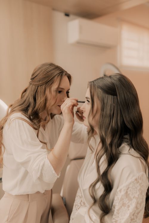 Free A makeup artist applies eye makeup to a woman in a warmly lit room, creating a serene atmosphere. Stock Photo