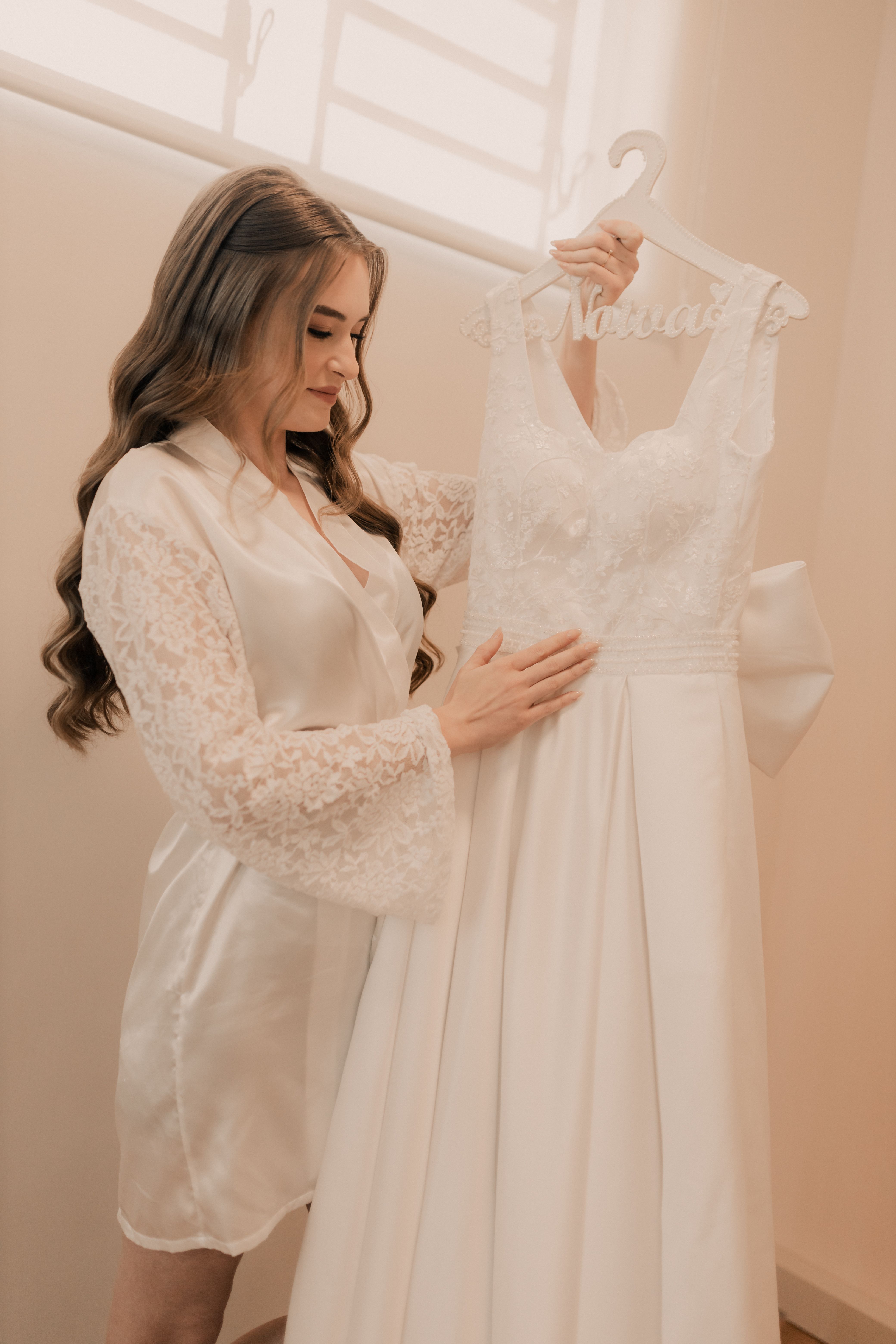 Bride in satin robe admiring her elegant wedding dress in a softly lit room.