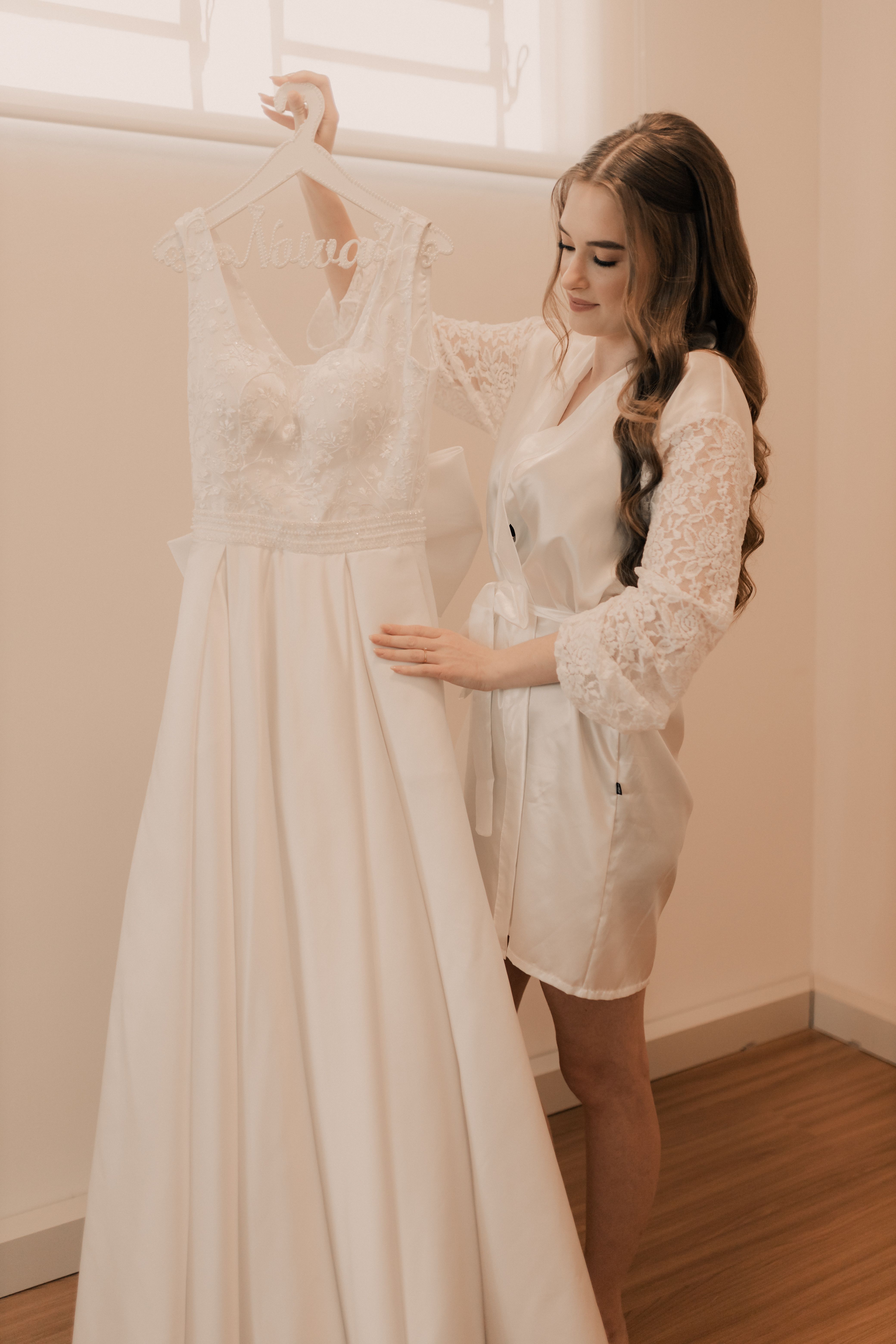 A bride admires her elegant white wedding gown, capturing a serene pre-wedding moment indoors.