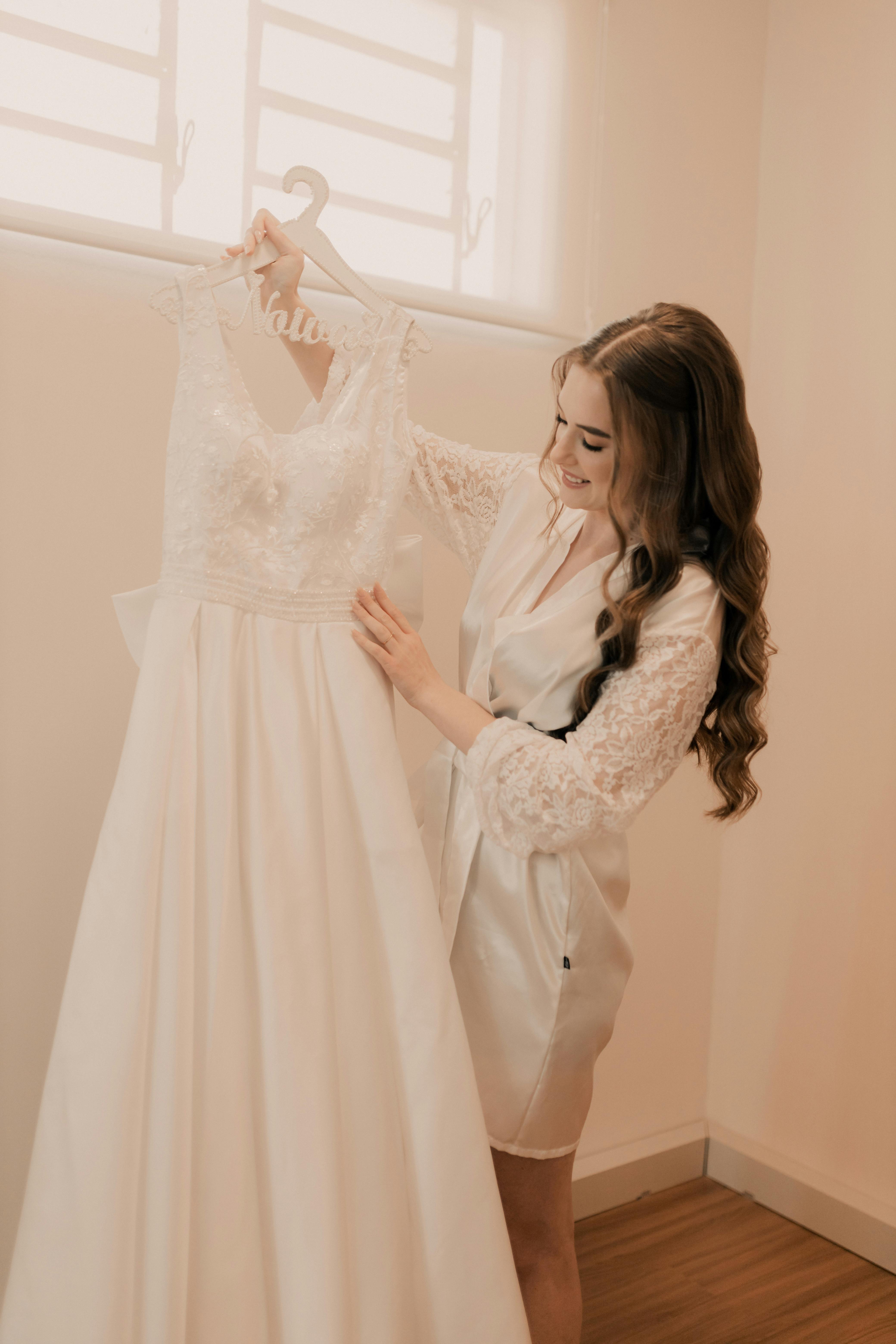 A bride joyfully looks at her white wedding dress in a sunlit room, capturing a moment of pre-wedding excitement.