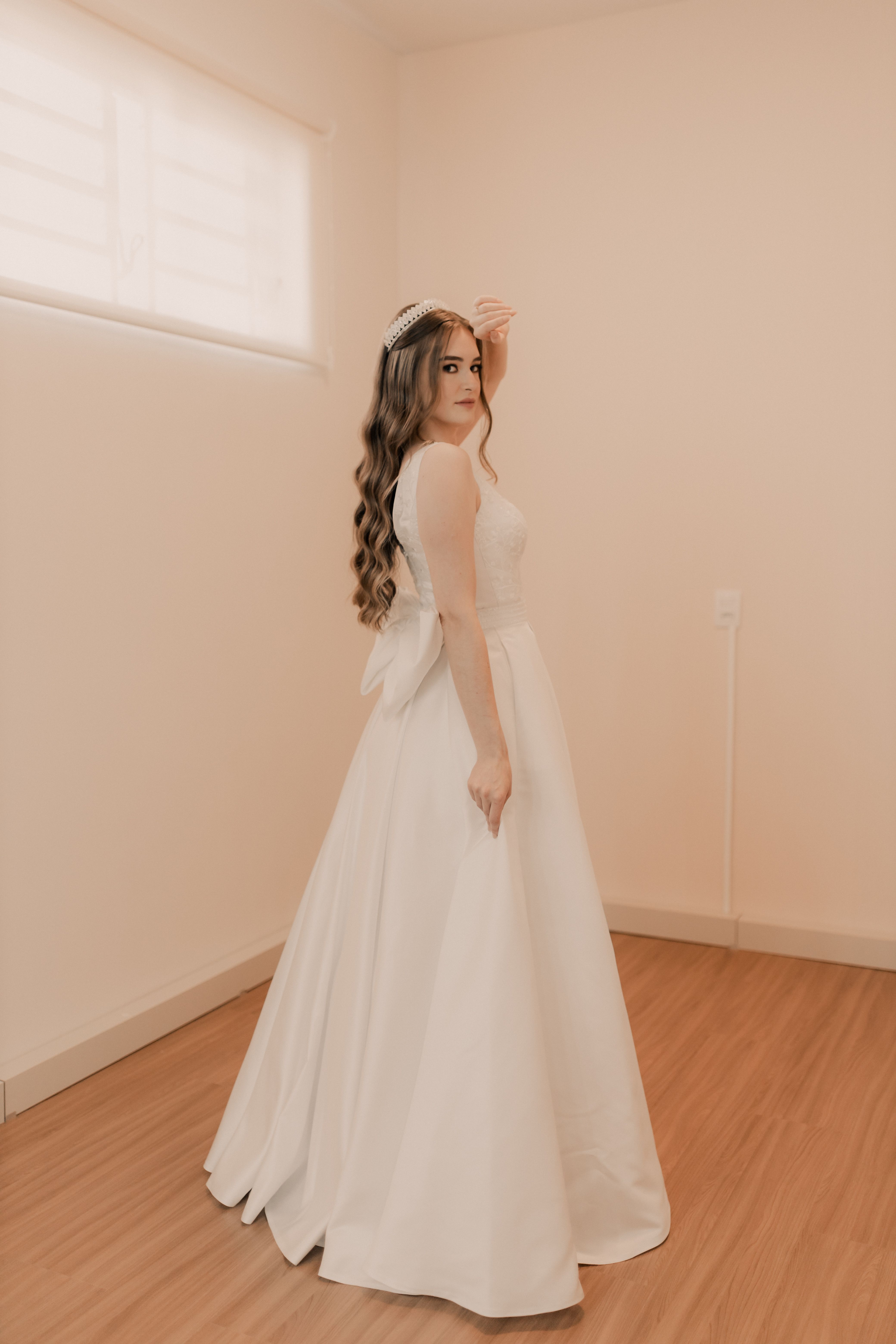 Bride in elegant white dress standing in a minimalist room with natural light from window.