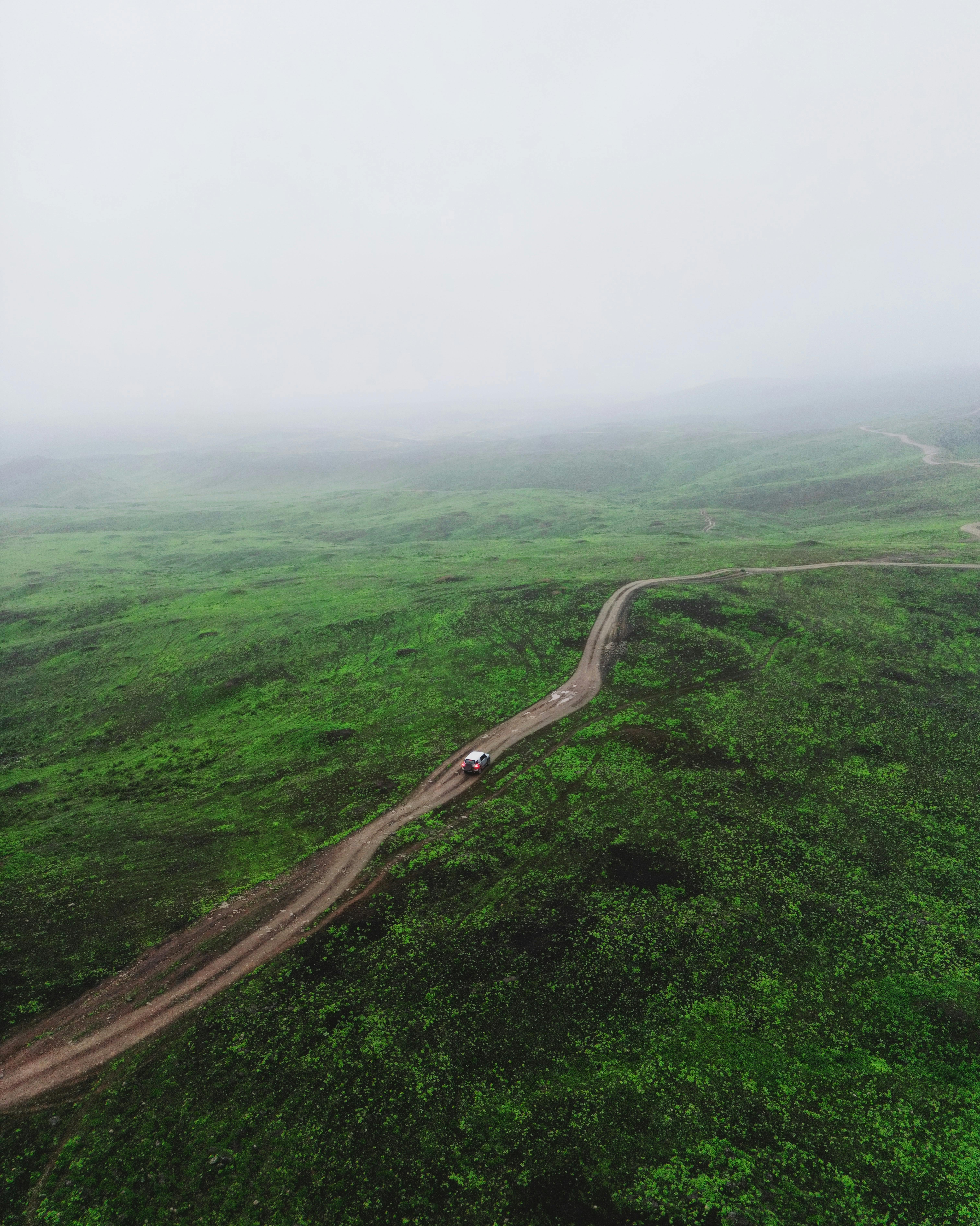 Aerial shot of a dirt road cutting through lush green hills in Oman, enveloped in mist.
