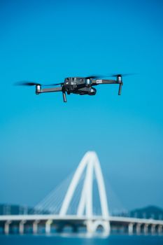 A drone hovers near a sleek arch bridge against a bright blue sky, showcasing technology with architectural beauty.