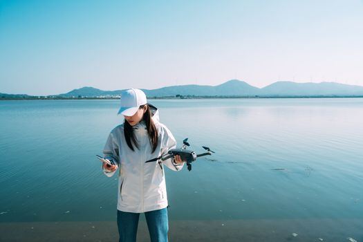 Young woman operating a drone by a tranquil lake with mountains in the background, on a clear day.