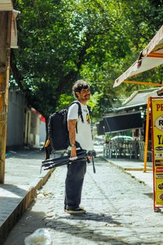 Photographer with gear standing on a vibrant city street under green trees.