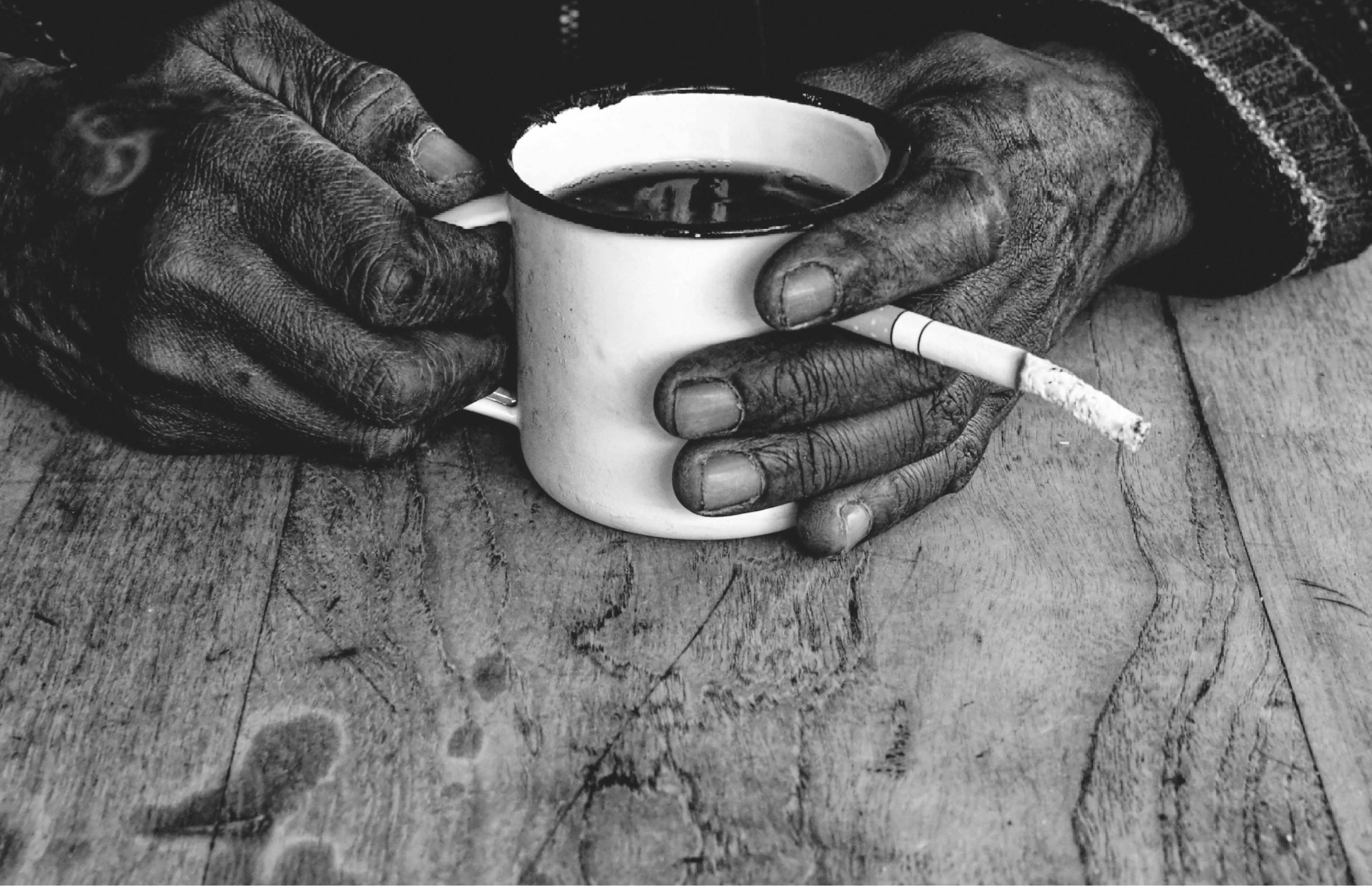 Close-up of elderly hands holding a coffee mug and cigarette, black and white.