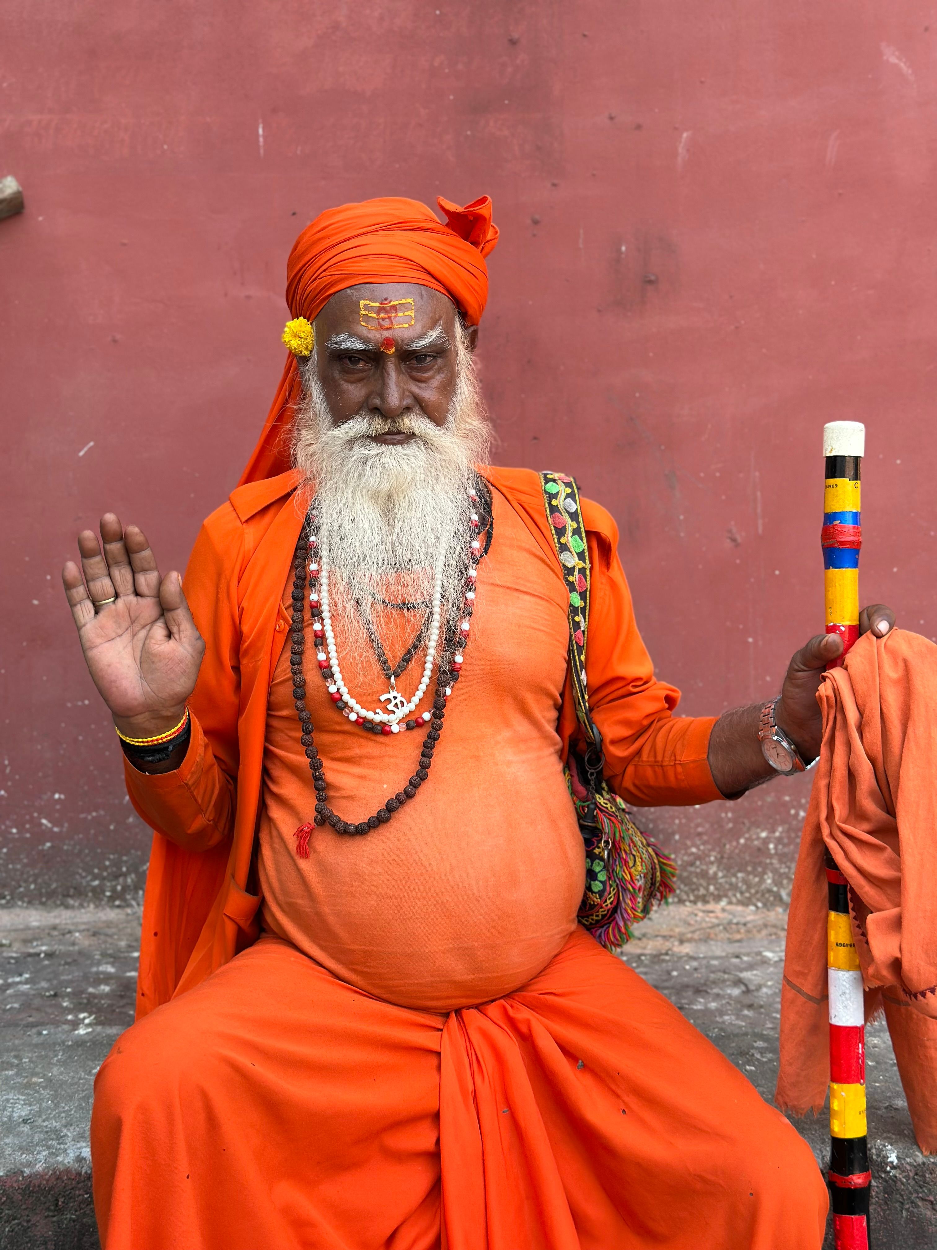 Elderly Sadhu dressed in vibrant orange robes meditating against a wall.