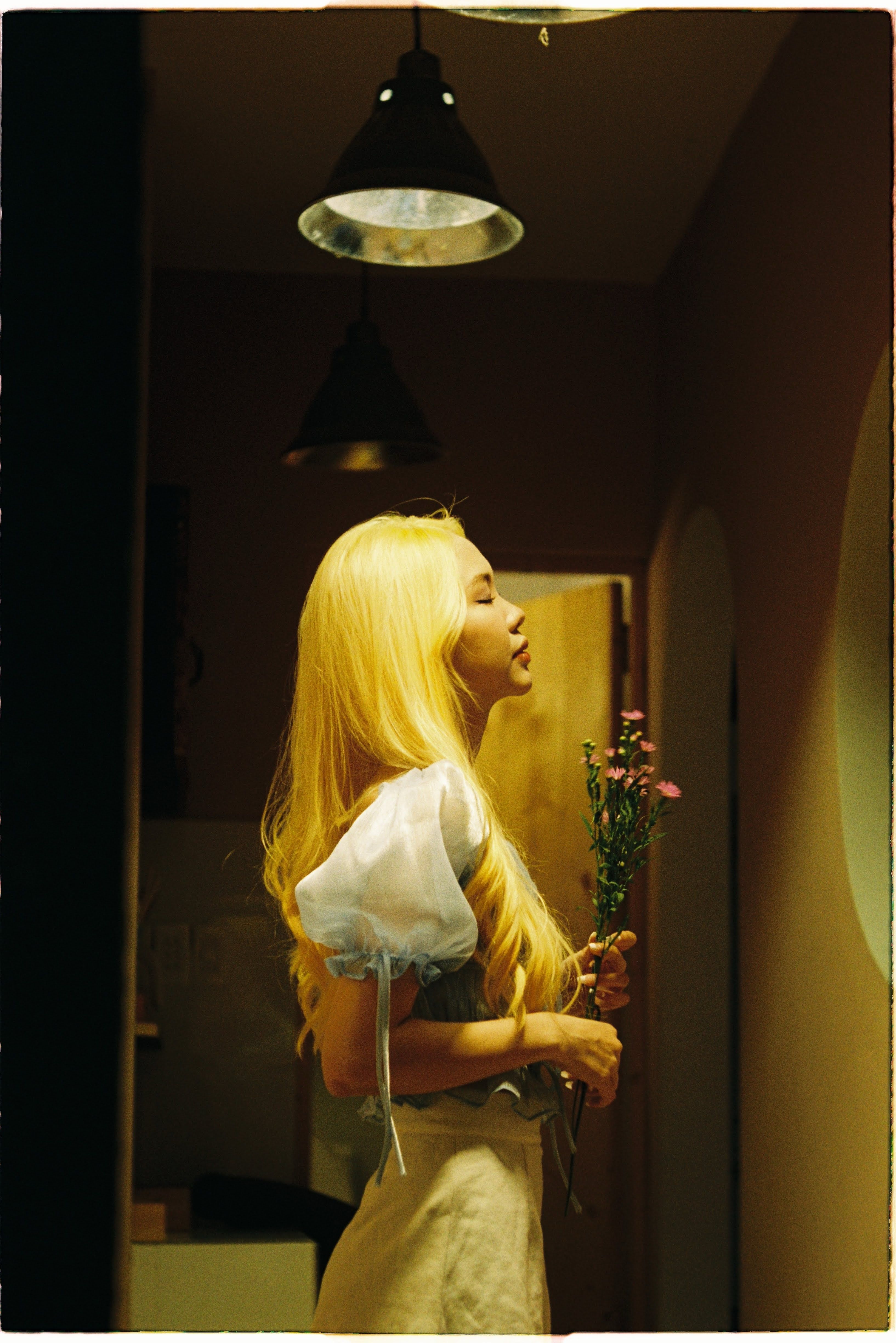 Woman with long blond hair holding flowers indoors under warm lighting.