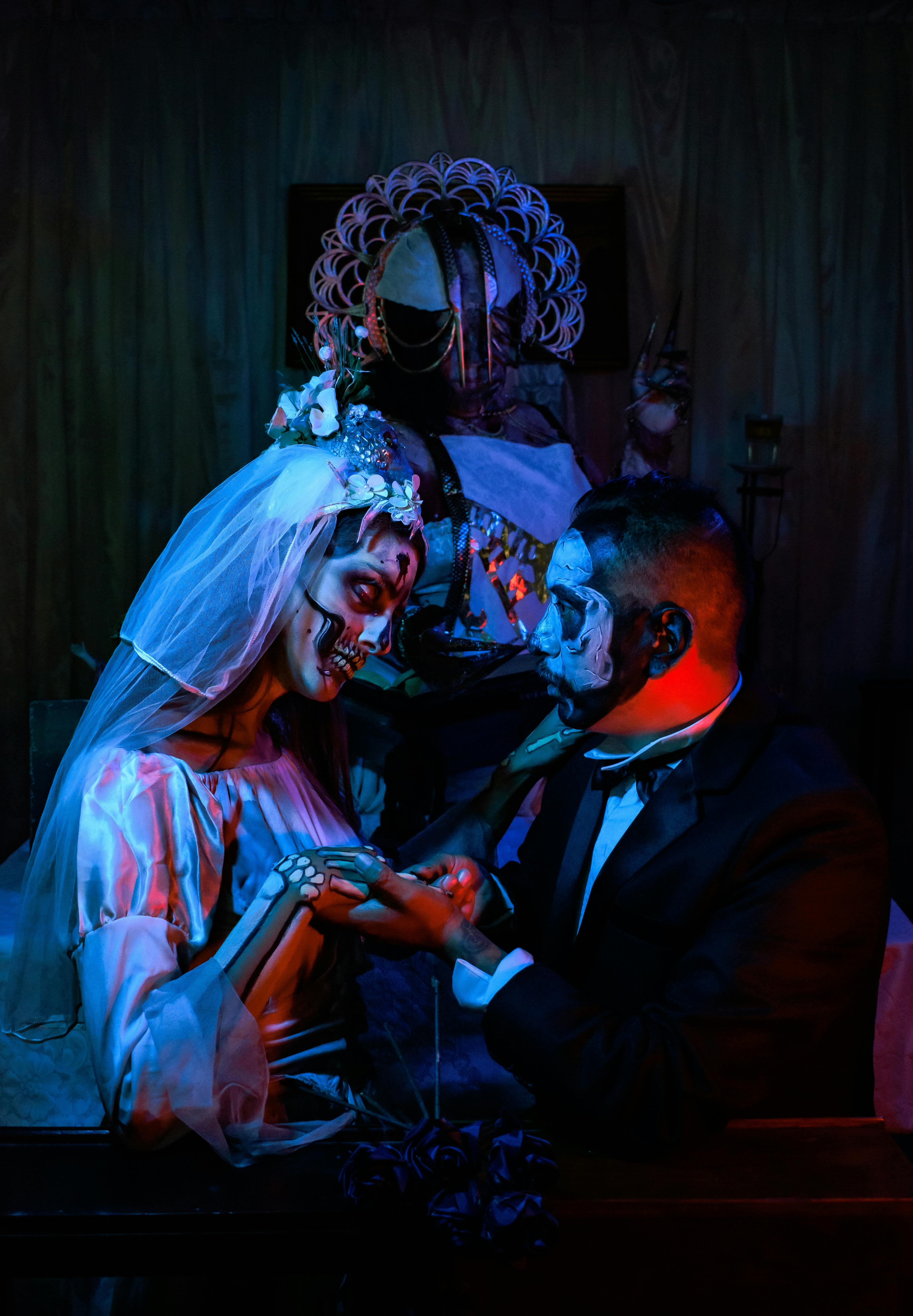 A couple dressed in Día de Muertos attire holds hands in a dark, dramatic setting.
