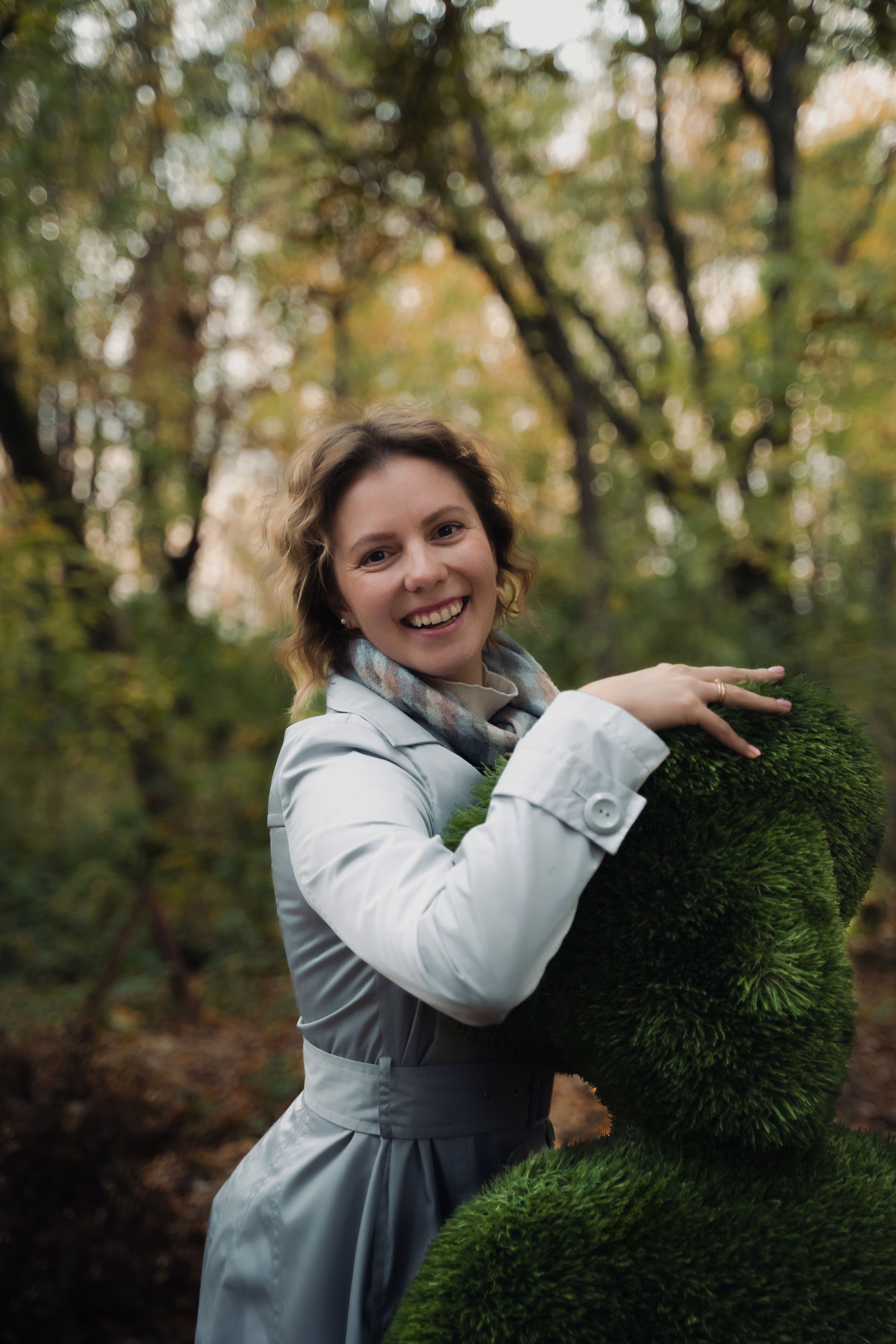 Woman in a light coat embracing a green topiary in a lush forest setting.