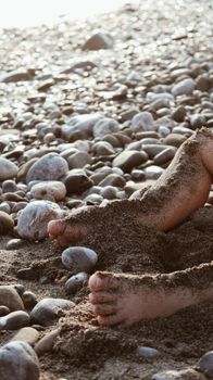 Sandy feet of a child buried in pebbles on a sunlit beach.