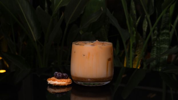 A refreshing iced coffee with cookie and fruit on black surface surrounded by green plants.