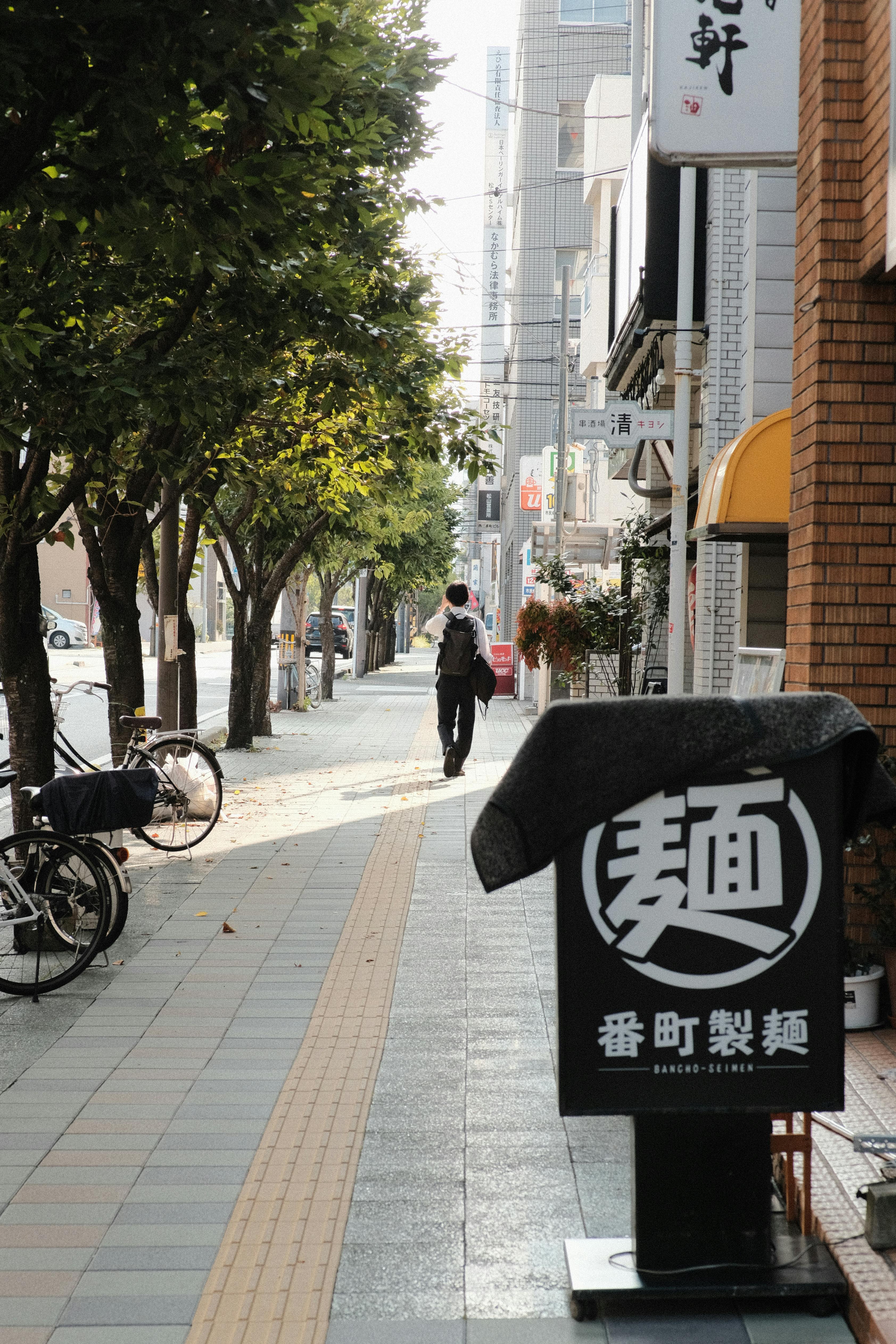 City sidewalk scene with cyclist, trees, and a noodle shop sign in soft daylight.