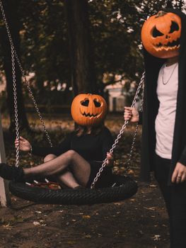Creative Halloween photo with two people wearing pumpkin masks, one sitting on a swing outdoors.