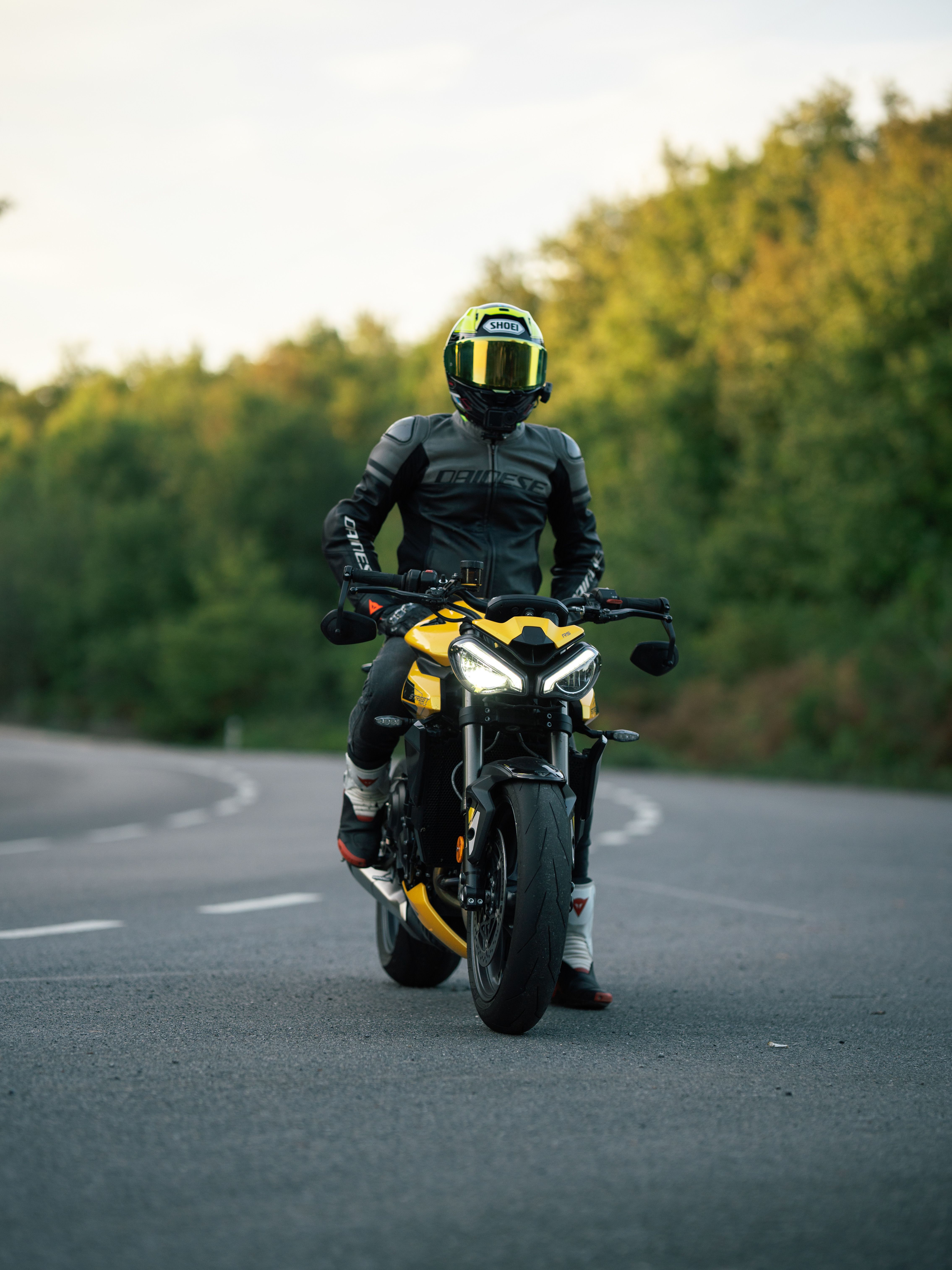 Person riding a yellow Triumph motorcycle on a winding road with green foliage.