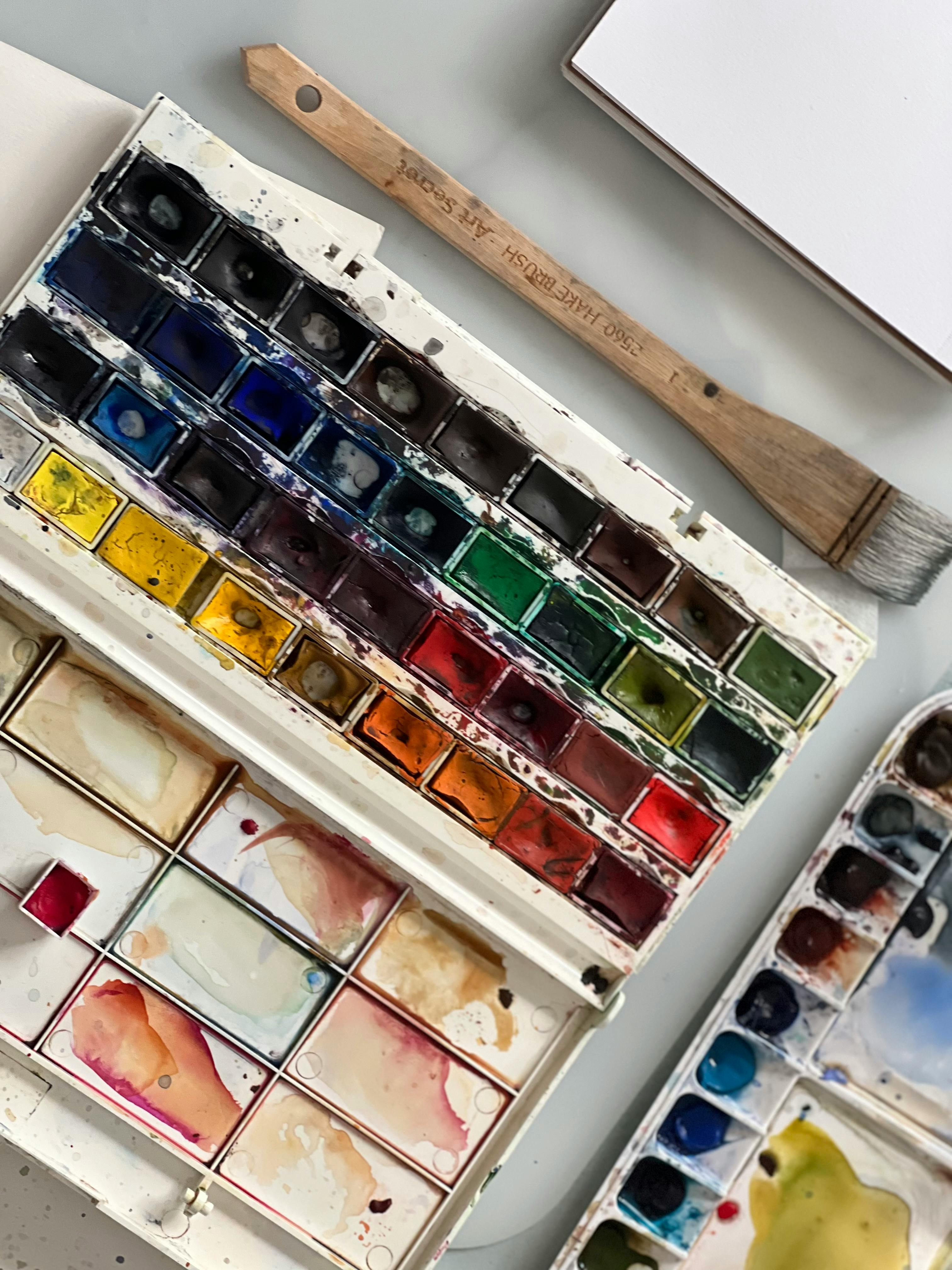 A vibrant watercolor paint set with a wooden paintbrush on a desk.