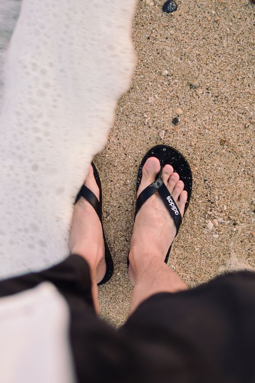 Free Bare feet in black flip flops by the ocean on a sandy beach. Stock Photo
