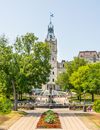 People By A Fountain In The Park With A  Walkway Surrounded With Trees And Flowers With In Front Of A Building With A Tower