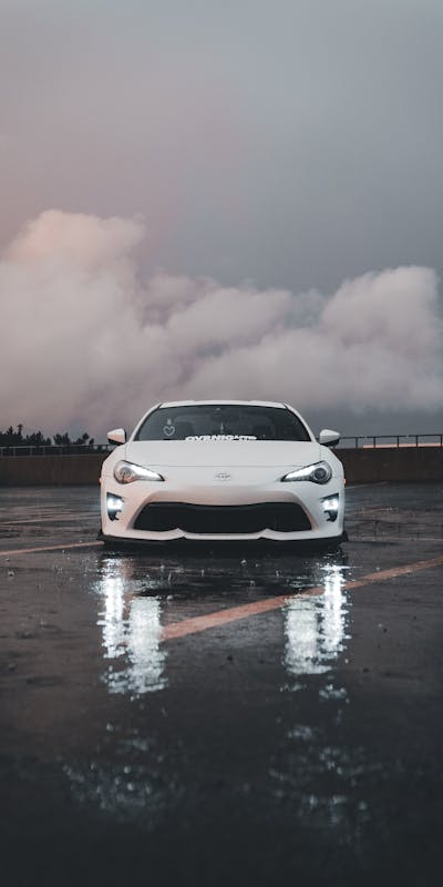 A white sports car parked on a wet rooftop, with reflections on the ground and a cloudy sky in the background.