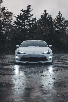 Front view of a white sports car on a rainy day in a forested area, showcasing sleek design and wet reflections.