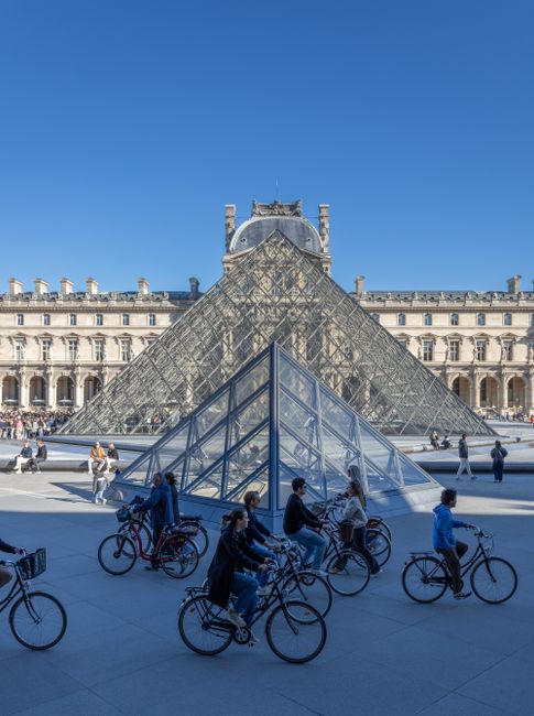 Cyclists pass by the iconic Louvre Pyramid in Paris under a clear blue sky.