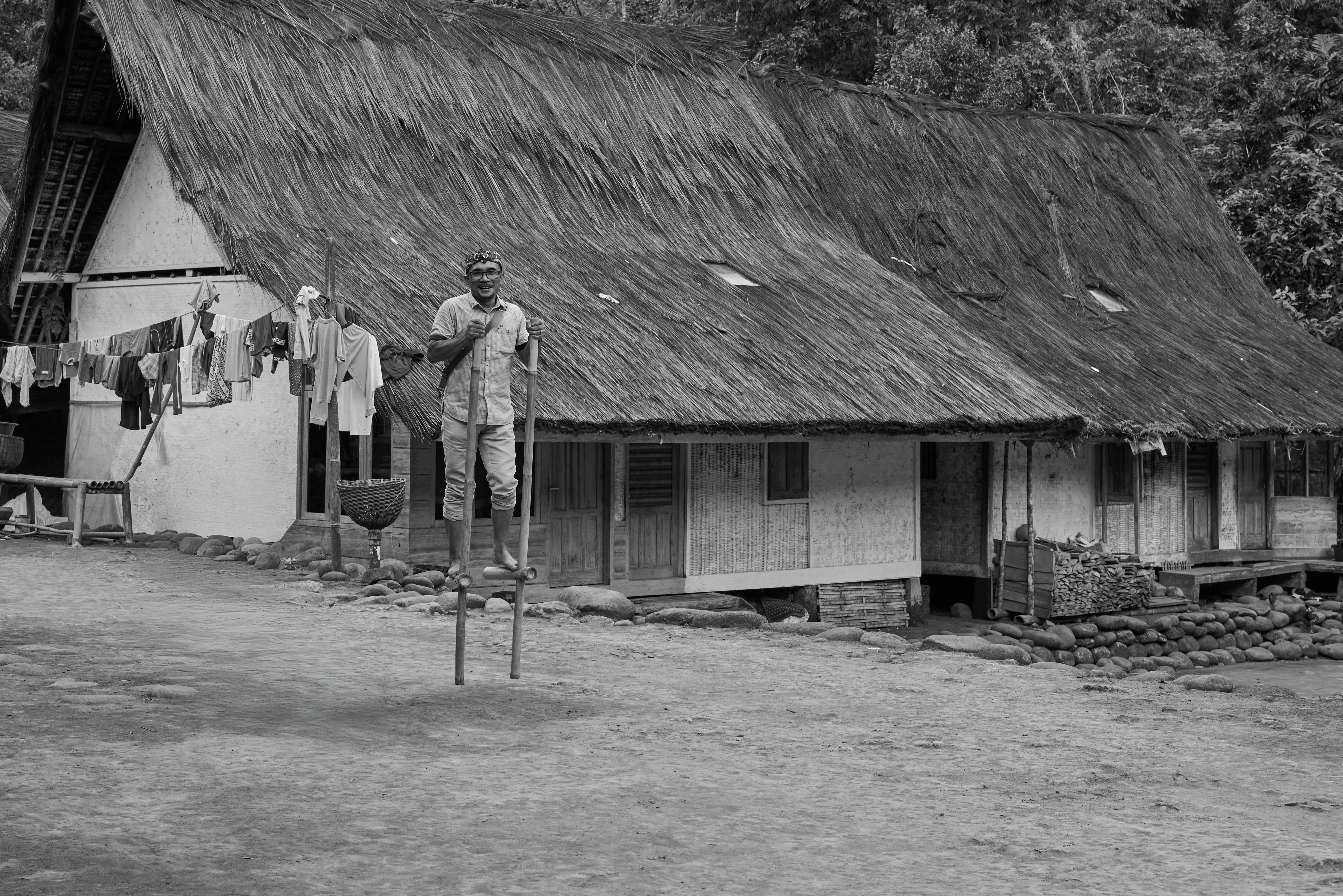 Black and white photo of a man on stilts in front of a thatched-roof house.