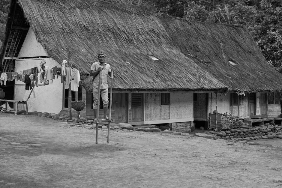 Black and white photo of a man on stilts in front of a thatched-roof house.