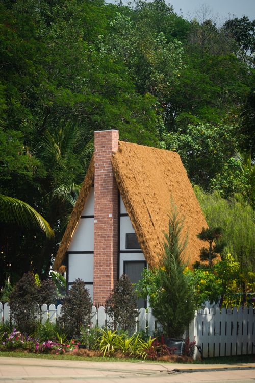 Free A picturesque A-frame house with a thatched roof surrounded by lush greenery and a white picket fence. Stock Photo