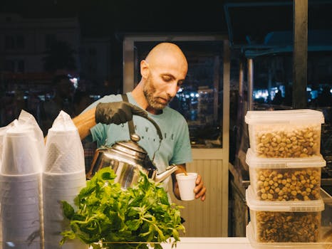 Bald street vendor wearing gloves pouring mint tea into a cup at a night market stall.